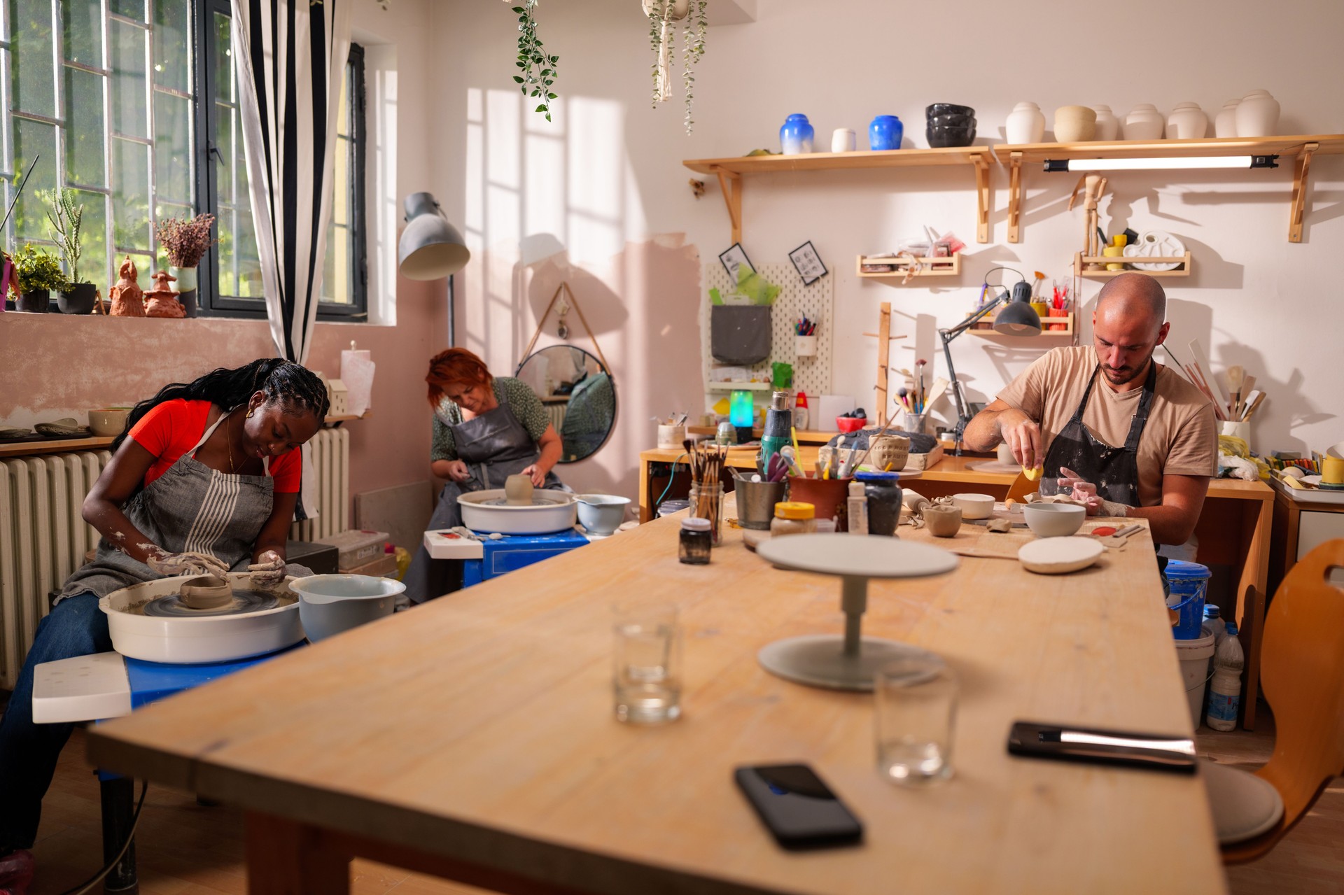 Three artisans, two women and a man, are working with clay in a pottery workshop, using pottery wheels and tools, surrounded by finished and unfinished ceramic pieces